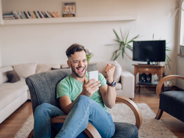 Mann sitzt mit Handy in der Hand auf Sessel und freut sich | WestLotto Gewinner Rubbellos Diamant 7