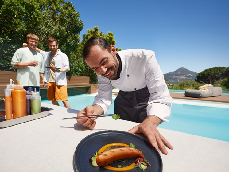 Koch steht vor einem Pool und verziert eine Rostbratwurst | Eurojackpot