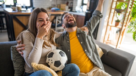 Eine Frau und ein Mann sitzen mit einem Fußball auf dem Sofa und jubeln - WestLotto TOTO Sondersauslosung