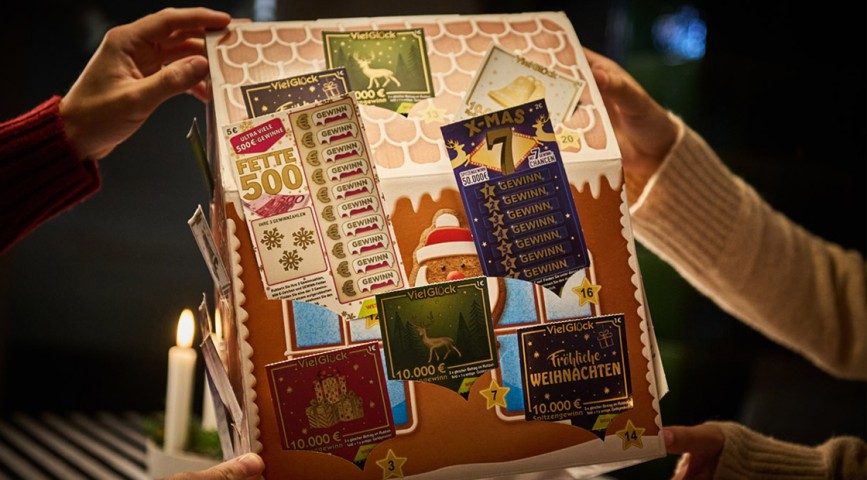 Vier Hände halten einen Rubbellos-Adventskalender in Lebkuchenform über einem Tisch - WestLotto