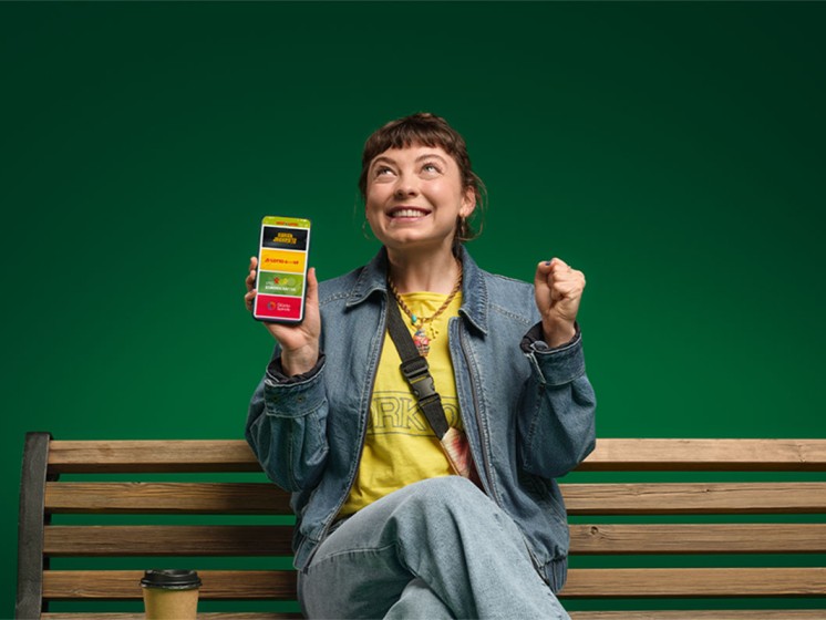 Frau sitzt auf einer Parkbank und hat ein Smartphone in der Hand, auf dem die WestLotto-App geöffnet ist. | WestLotto