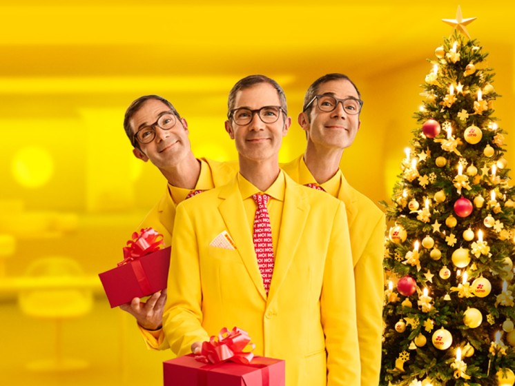 Mann steht vor Tannenbaum, hält Geschenk in der Hand und grinst