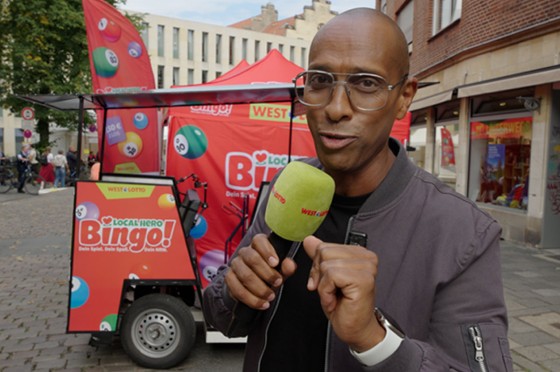 Moderator Amiaz Habtu mit Mikrofon in der Hand steht vor einer Rikscha - WestLotto Local Hero Bingo