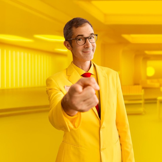 Mann mit gelbem Anzug steht vor gelbem Hintergrund und zeigt mit Finger auf Betrachter - WestLotto Lotto-SuperDing