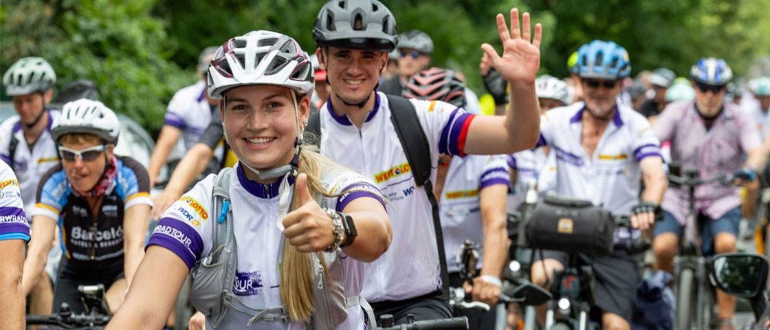 Mehrere Radfahrer auf einer Straße. Zwei von Ihnen winken in die Kamera. NRW-Radtour WestLotto