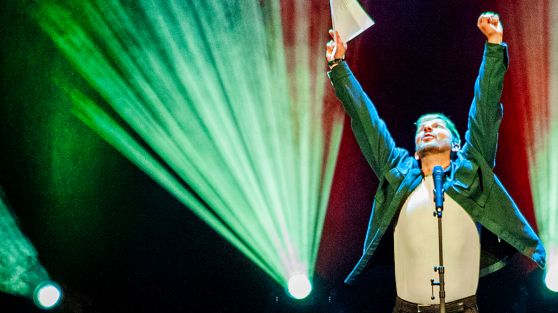 Poetry-Slammer steht auf Bühne, streckt Arme in die Höhe und schaut nach oben - WestLotto Poetry-Slam